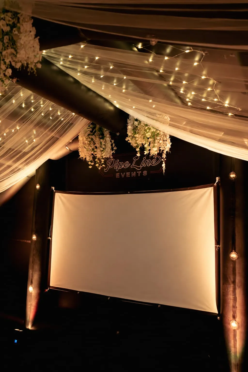 Milestone birthday setup with white draping, fairy lights, hanging florals and a welcome screen